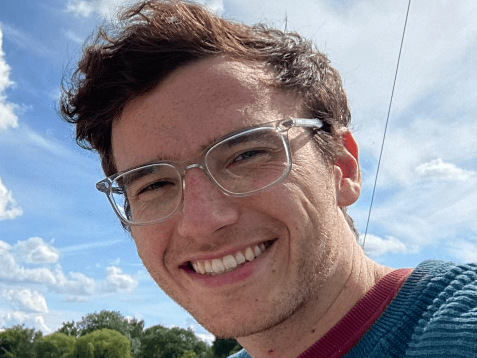 Junger Mann mit Brille und Pullover lächelt am Ufer eines Gewässers, im Hintergrund Bäume und wolkiger Himmel.