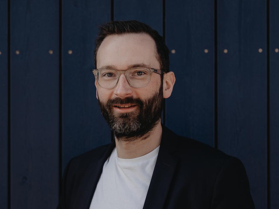 Mann mit Brille und Bart steht vor einer blauen Wand, trägt einen schwarzen Blazer und ein weißes T-Shirt.
