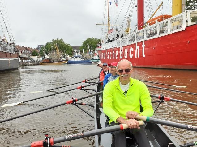 Rudergruppe in einem Boot auf einem Wasserweg, im Hintergrund ein rot gestrichenes Schiff und Hafenansicht.