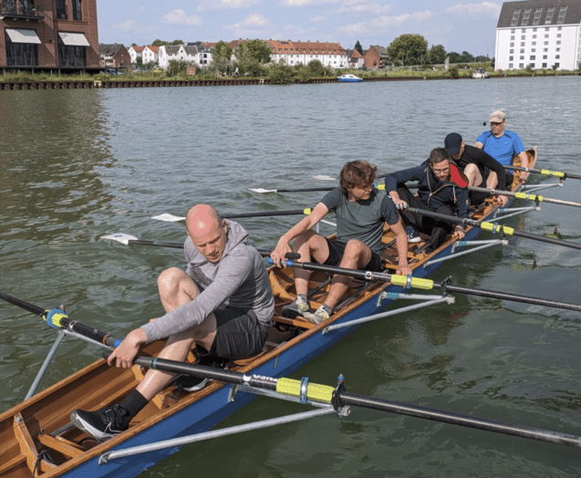 Sechs Ruderer in einem Boot auf einem Gewässer, während sie synchron rudern und im Hintergrund Ufergebäude sichtbar sind.