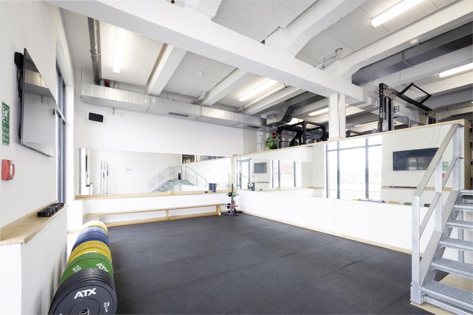 Blick in einen modernen Trainingsraum mit Spiegeln, Gewichtsscheiben und Treppe im Hintergrund.