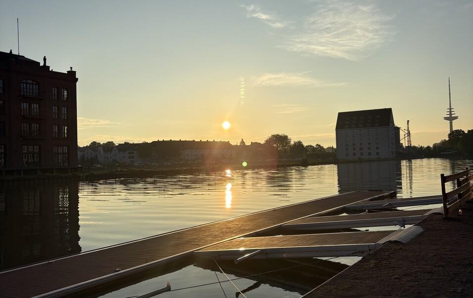 Sonnenuntergang &uuml;ber einem ruhigen Gew&auml;sser mit Holzsteg und historischen Geb&auml;uden im Hintergrund.