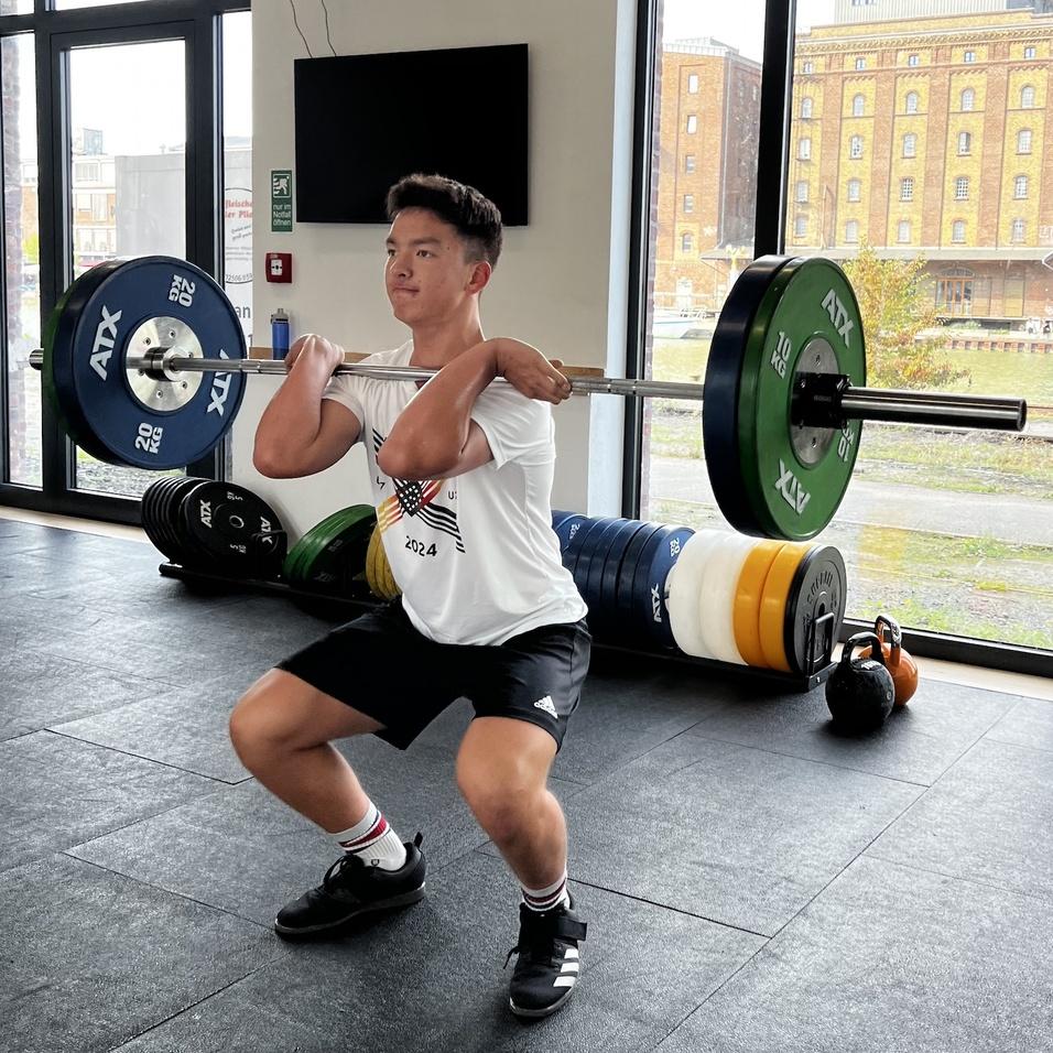Junger Mann hebt eine Langhantel in einer Fitnessstudio-Umgebung, große Fenster im Hintergrund.