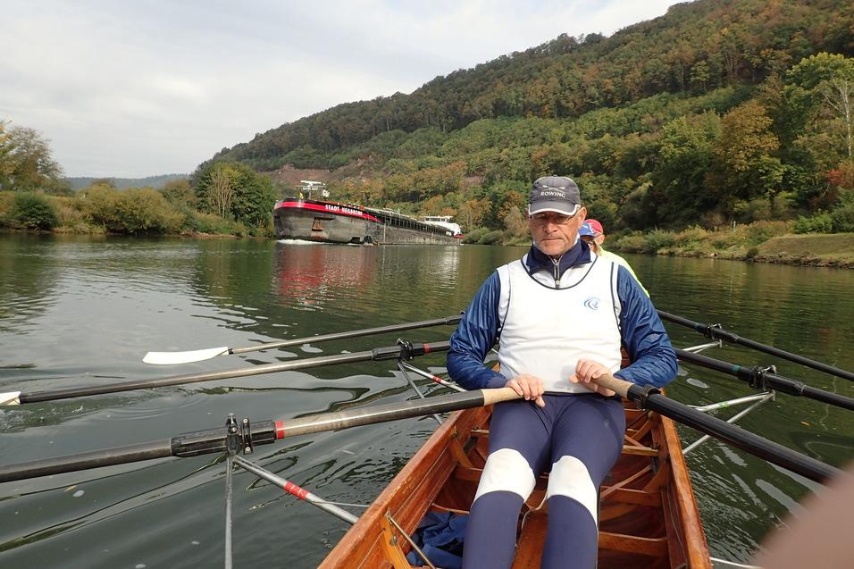 Ruderer sitzt im Boot am Fluss, während ein Frachtschiff im Hintergrund vorbeifährt. Bunte Baumreihe am Ufer.