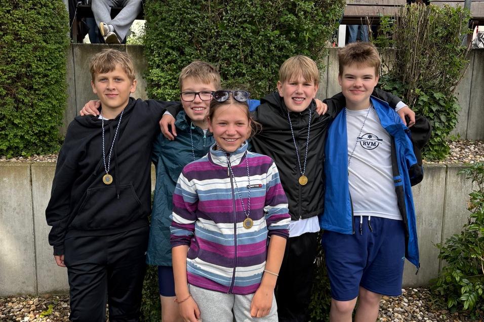 F&uuml;nf Kinder stehen l&auml;chelnd zusammen, tragen Medaillen und Freizeitkleidung vor einer gr&uuml;nen Hecke.