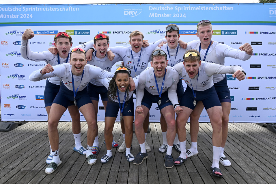 Gruppe von neun jubelnden Athleten in Bootskleidung mit Medaillen vor einem Banner der Deutschen Sprintmeisterschaften.