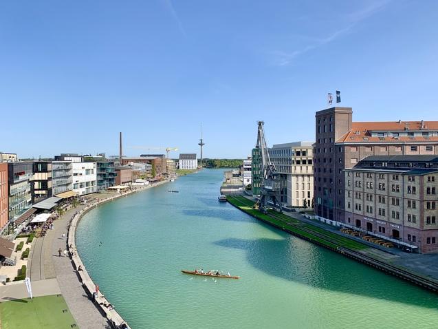 Blick auf einen klaren Kanal mit modernem Wohn- und Gewerbebau sowie einem Kanu mit Sportlern auf dem Wasser.