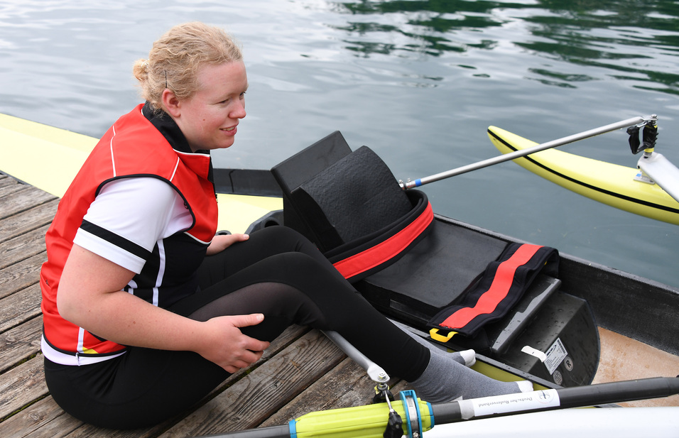 Junge Frau in roter Schwimmweste sitzt auf einem Ruderboot an einem Steg, mit Ruder und Boot im Hintergrund.