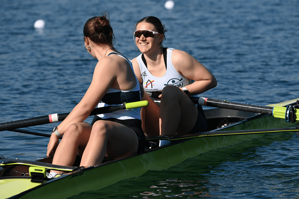 Zwei Ruderinnen sitzen in einem Doppelboot und l&auml;cheln auf einem Gew&auml;sser bei sonnigem Wetter.